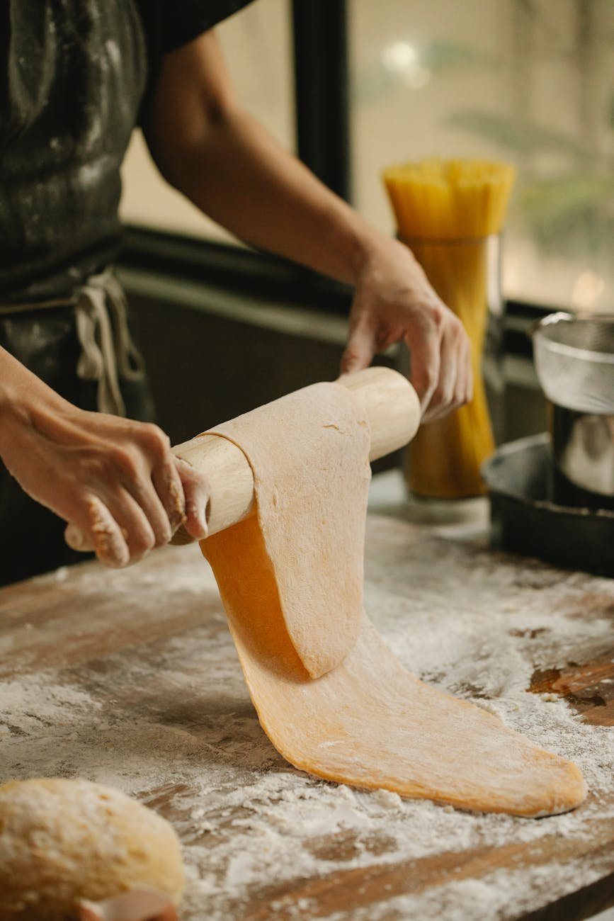 unrecognizable female rolling pasta dough on floury table at home